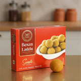 Box of Besan Laddu sweets with a single laddu on a reflective surface.