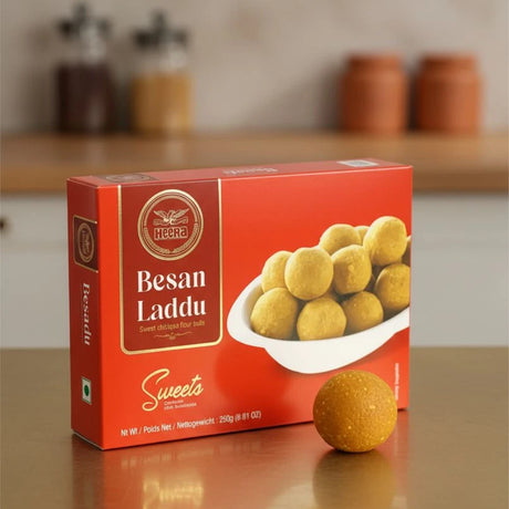 Box of Besan Laddu sweets with a single laddu on a reflective surface.